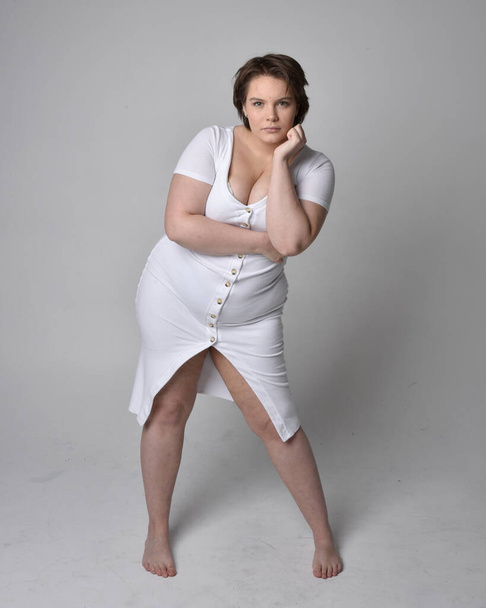 Full length portrait of young plus sized woman with short brunette hair,  wearing a tight white body con dress,   standing walking  posewith light studio background. - Photo, Image