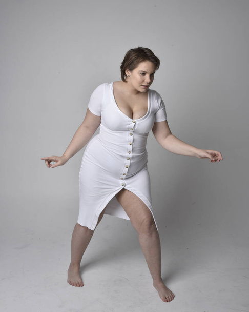 Full length portrait of young plus sized woman with short brunette hair,  wearing a tight white body con dress,   standing walking  posewith light studio background. - Foto, afbeelding