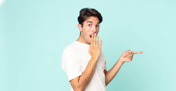 hispanic handsome man feeling happy, shocked and surprised, covering mouth with hand and pointing to lateral copy space - Фото, зображення