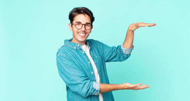 hispanic handsome man smiling, feeling happy, positive and satisfied, holding or showing object or concept on copy space - 写真・画像