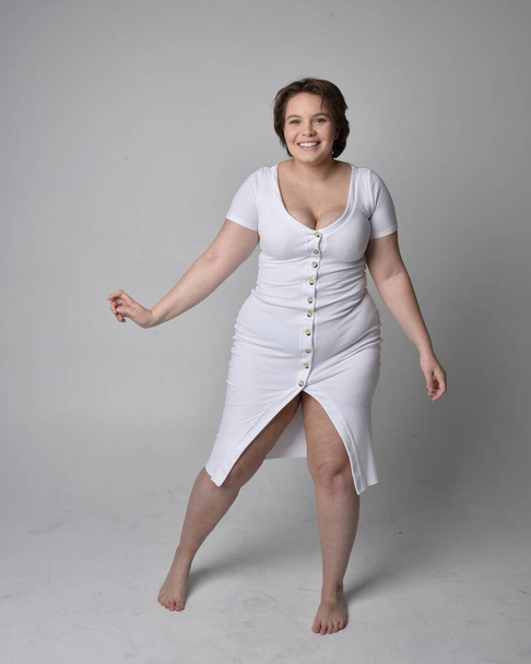Full length portrait of young plus sized woman with short brunette hair,  wearing a tight white body con dress,   standing walking  posewith light studio background. - Foto, Imagen