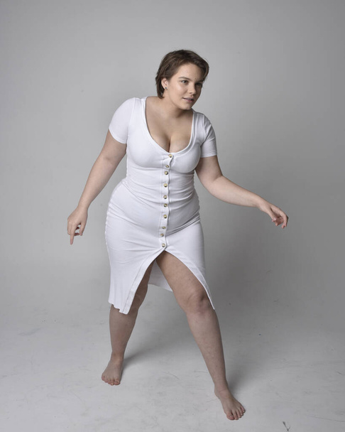 Full length portrait of young plus size woman with short brunette hair, wearing a tight white body con dress, standing walking posewith light studio background. - Foto, afbeelding