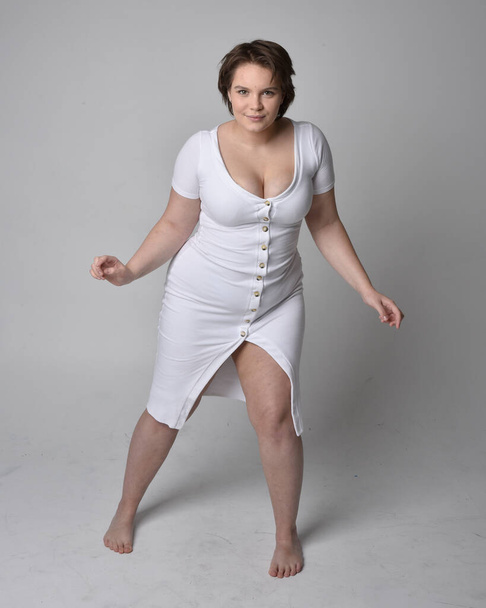 Портрет молодої плюс розмір жінки з коротким волоссям брюнетки, одягнений у вузьку білу сукню конюшини, що стоїть на світлому фоні студії
. - Фото, зображення