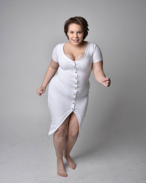 Full length portrait of young plus sized woman with short brunette hair,  wearing a tight white body con dress,   standing walking  posewith light studio background. - Foto, Bild