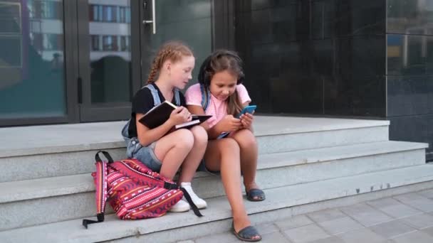  Dos chicas con mochila sentarse y leer libro con teléfono - Imágenes, Vídeo