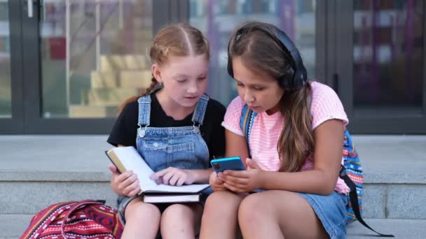  Zwei Mädchen mit Rucksack sitzen und lesen Buch mit Handy - Filmmaterial, Video