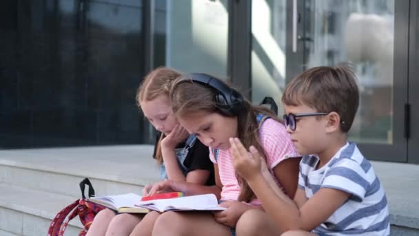  Трое детей с рюкзаком сидят и читают книги, играют в телефон - Кадры, видео
