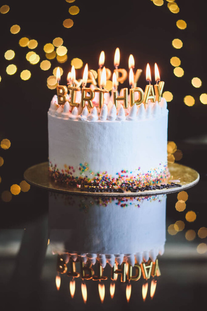Verjaardagstaart met kaarsen, slinger met felle bokeh lichten op de achtergrond. De witte taart is versierd met gekleurde hagelslag en staat op een reflecterend oppervlak. - Foto, afbeelding