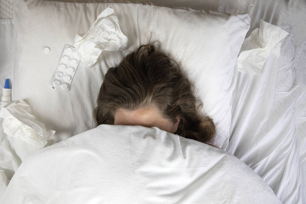 A sick young woman in bed, lying with her head under the blankets and a pile of tissues next to her top view. Fever staying home from work. white sheets and pillow - Photo, Image