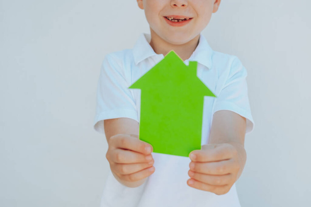 Zamknij dłonie dzieci trzymając dom z zielonego papieru, skup się na uśmiechu chłopca. na białym tle. koncepcja ekologicznych domów. Wysokiej jakości zdjęcie - Zdjęcie, obraz