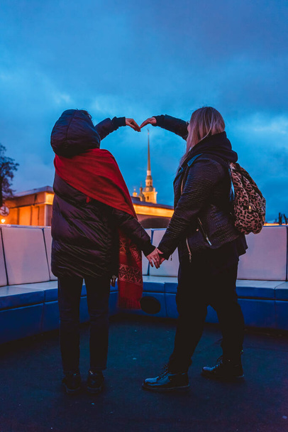 two girls form a heart from their hands around the Peter and Paul Cathedral, standing on the deck of a boat passing around the Peter and Paul fortress in the center of St. Petersburg at night - Photo, Image