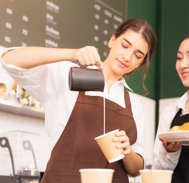 Beautiful caucasian barista women fill white milk into takeaway hot coffee cup at coffee bar in cafe coffee shop. Barista work at coffee bar and food service business - Foto, afbeelding