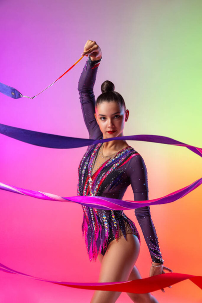 Een mooie jonge vrouwelijke ritmische gymnaste poseren met helder lint in haar hand geïsoleerd op veelkleurige neon achtergrond. - Foto, afbeelding