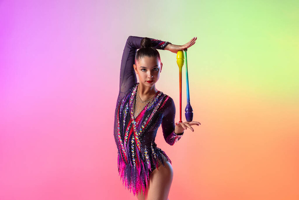 Una gimnasta rítmica femenina con clubes posando aislados sobre fondo de neón multicolor. Retrato de media longitud - Foto, Imagen