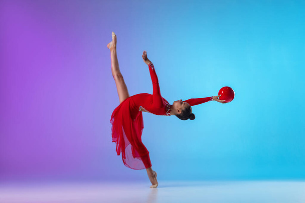 Jong meisje, ritmische turnster in rode jurk met bal in beweging en actie geïsoleerd op paars blauw gradiënt neon achtergrond. - Foto, afbeelding