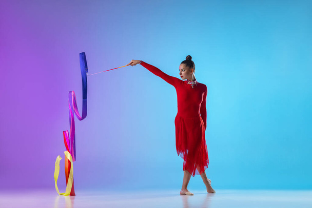 Porträt einer rhythmischen Turnerin im roten Kleid, die mit einem hellen Band auf einem abfallenden Neon-Hintergrund trainiert. - Foto, Bild