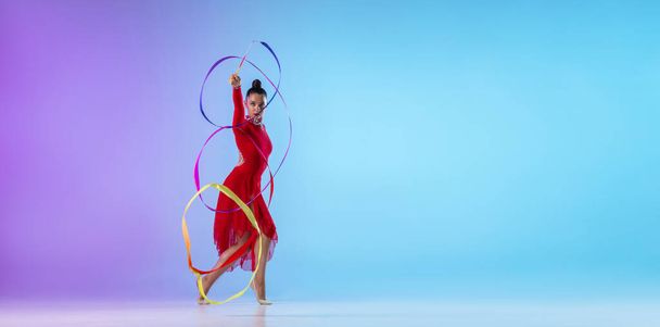 Een flyer. Portret van vrouwelijke ritmische turnster in rode jurk oefenen met helder lint geïsoleerd op gradiënt neon achtergrond. - Foto, afbeelding