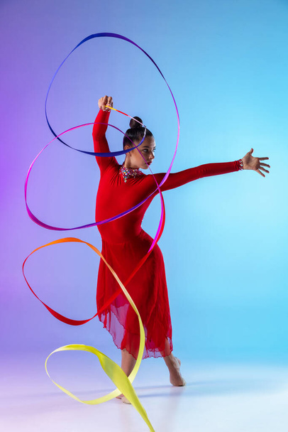 Young professional female rhythmic gymnast exercising with bright ribbon isolated on gradient neon background. - Φωτογραφία, εικόνα