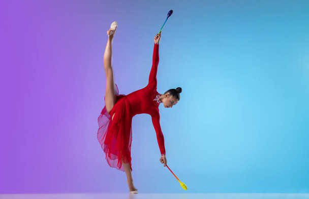 Young girl, rhythmic gymnast in red dress exercising with clubs isolated on gradient neon background. - Foto, Imagem