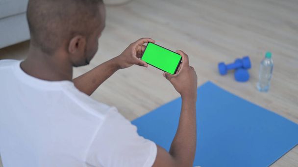 Vista trasera del hombre africano mirando el teléfono inteligente con pantalla clave de croma en la estera de yoga - Foto, Imagen