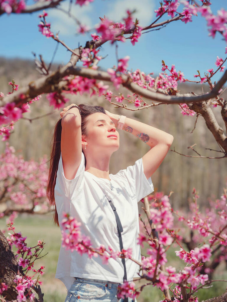 beautiful woman with dark hair in white t-shirt among blossoming peach trees in park. Looking at camera. Spring season. - 写真・画像
