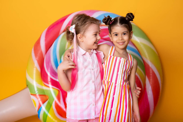 two happy little girls in colorful dress laughing hugging having fun on yellow background with lollipop. - Photo, Image
