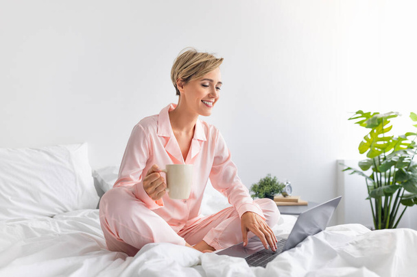 Szczęśliwa kobieta siedzi na łóżku używając komputera pijąc kawę - Zdjęcie, obraz