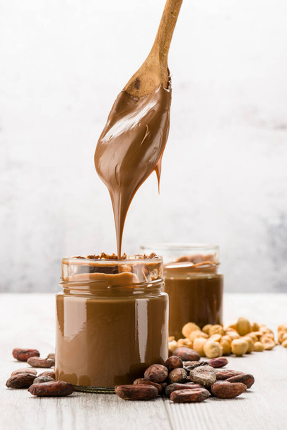 Chocolate cream in glass jar with cocoa beans, hazelnuts and dripping spoon, on wooden table - Foto, Bild