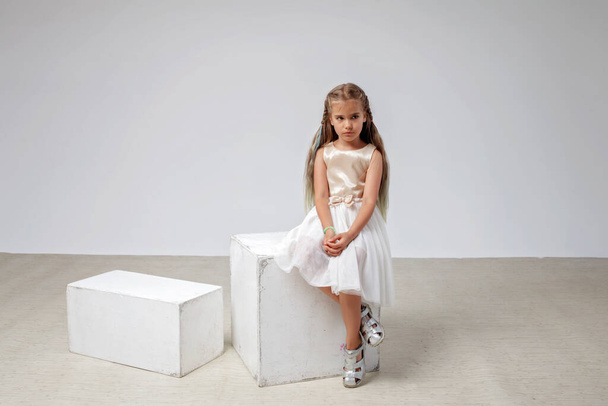 Sad girl with long hair in festive dress sits on square podium, white background, studio shot - Photo, Image