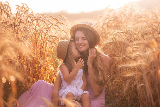 Close-up portrait of mother with daughter in straw hats in wheat field with dew drops. Young woman have fun with girl, tickle each other with golden ears of rye, laugh hug kiss each other. Countryside - Фото, зображення
