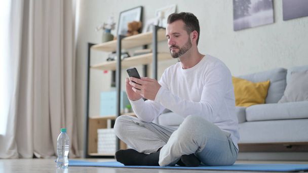 Homem adulto maduro usando Smartphone no tapete de ioga em casa - Foto, Imagem