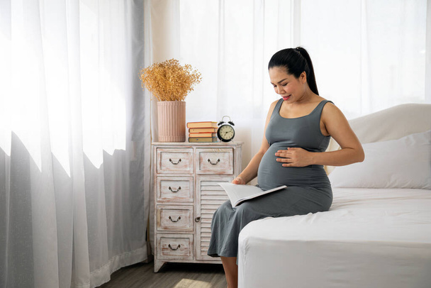 Happy Asia pregnant woman reading book and sitting on white bed. Relaxation give birth mother - Foto, Bild