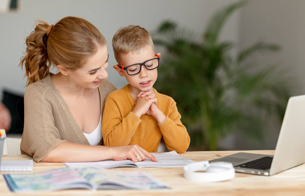 Összpontosított kis iskolás csinál gyakorlatokat a copybook, miközben tanul távolról laptopon keresztül otthon boldog anya ül a közelben, és támogatást nyújt - Fotó, kép