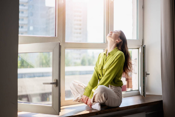 Beautiful young happy woman girl brunette in a green blouse sitting on the windowsill at home - Foto, afbeelding