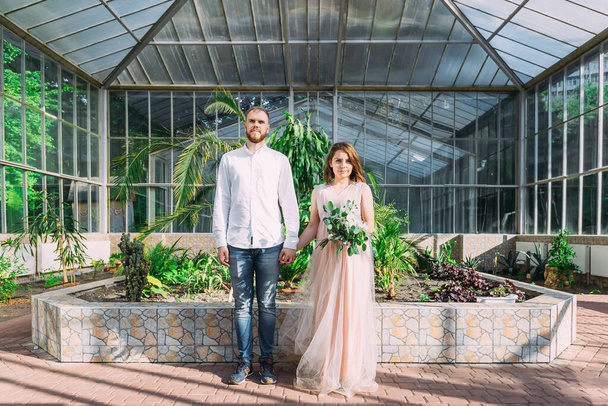 Gli sposi camminano nel giardino botanico vicino all'antica architettura di vetro, si aggrappano alle mani - Foto, immagini
