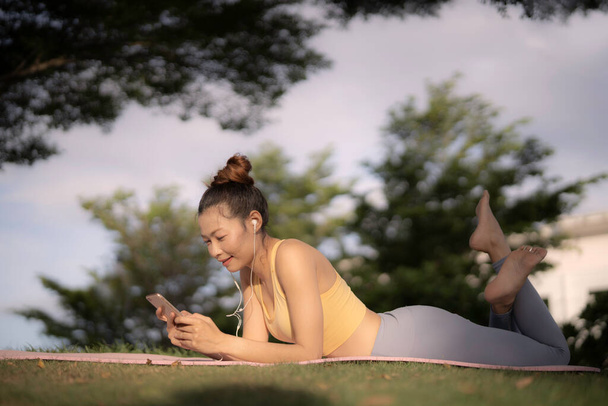 Una mujer asiática en sujetador deportivo amarillo y gris rezagado se tumbó en la estera de yoga y el suelo de hierba bajo un gran árbol después de hacer ejercicio sosteniendo el teléfono inteligente y escuchar música desde los auriculares con sonrisa. Relajante, concepto de mente curativa. - Foto, Imagen