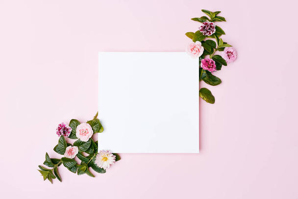 ピンクのパステルの背景に花で飾られた空白の紙のカード。コピースペースでのトップビューのモックアップ. - 写真・画像