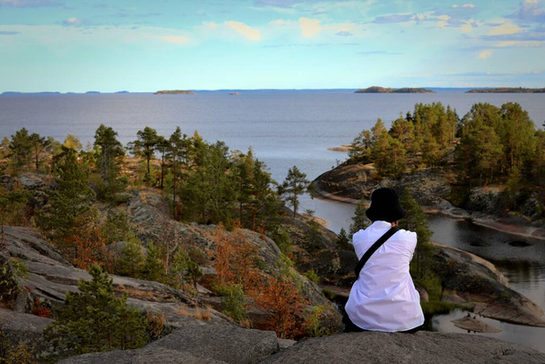dívka turista v bílé košili a černé panama sedí obdivuje výhled na skerries, skalnaté ostrovy jezera Ladoga v letním dni - Fotografie, Obrázek