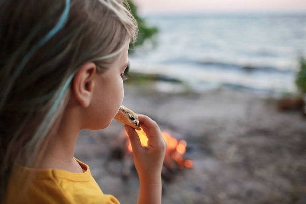 Girl roasting marshmallow to make smores over fire flame during camping, traditional travel food - Fotó, kép