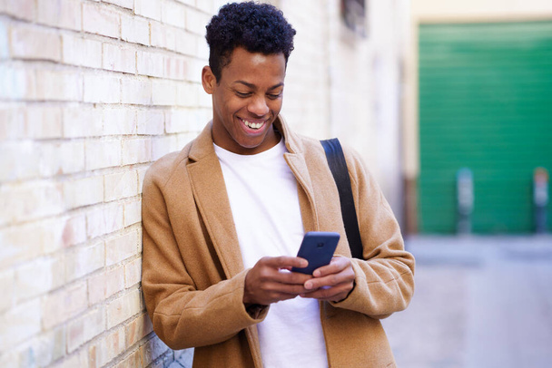 Jonge zwarte man typen op zijn smartphone leunend tegen een bakstenen muur in de straat. - Foto, afbeelding