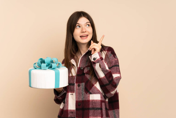 Young Ukrainian girl holding a big cake isolated on beige background intending to realizes the solution while lifting a finger up - Photo, Image
