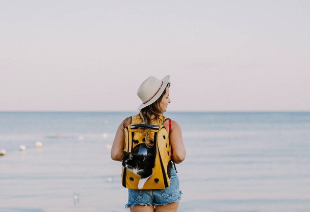 young woman with a cat in a backpack on the seashore. Travel concept with a pet. - Foto, immagini