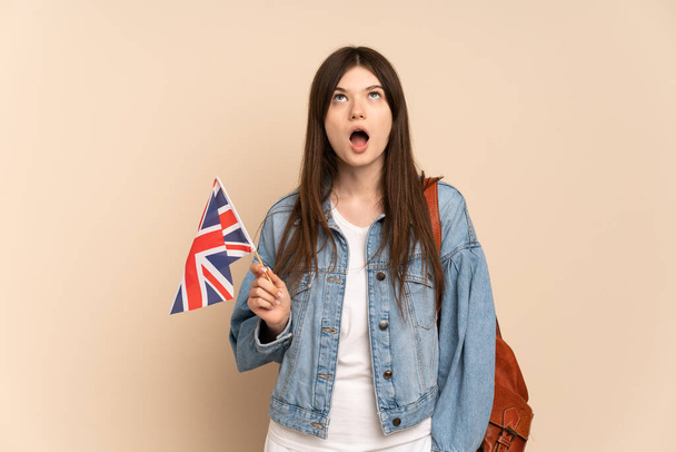 Ukraynalı genç bir kız bej arka planda izole edilmiş bir Birleşik Krallık bayrağını tutuyor yukarı bakıyor ve şaşırmış bir ifadeyle - Fotoğraf, Görsel