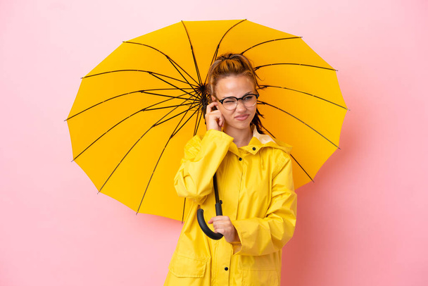Teenager rusky dívka s nepromokavý kabát a deštník izolované na růžovém pozadí frustrovaný a zakrývající uši - Fotografie, Obrázek