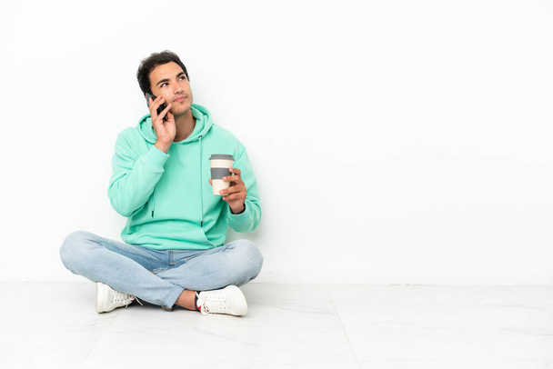 Кавказький вродливий чоловік сидить на підлозі, тримаючи каву, щоб забрати його і мобільний. - Фото, зображення