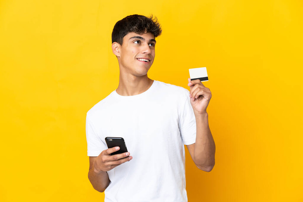Młody człowiek na odizolowanym żółtym tle zakupu za pomocą telefonu komórkowego z karty kredytowej podczas myślenia - Zdjęcie, obraz