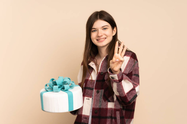 Joven ucraniana sosteniendo un gran pastel aislado sobre fondo beige feliz y contando tres con los dedos - Foto, Imagen