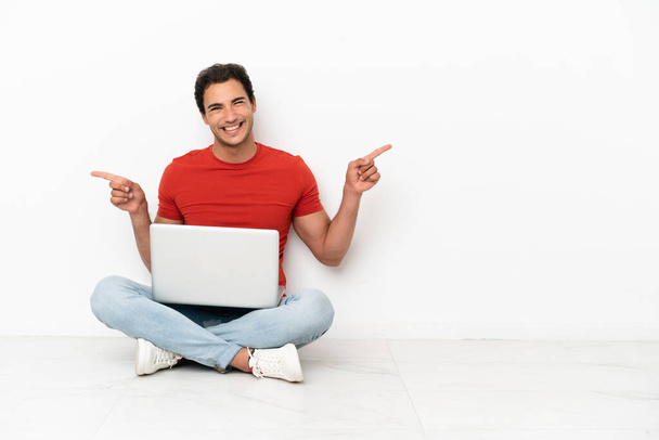Valkoihoinen komea mies kannettavan tietokoneen kanssa istuu lattialla osoittaen sormella laterals ja onnellinen - Valokuva, kuva