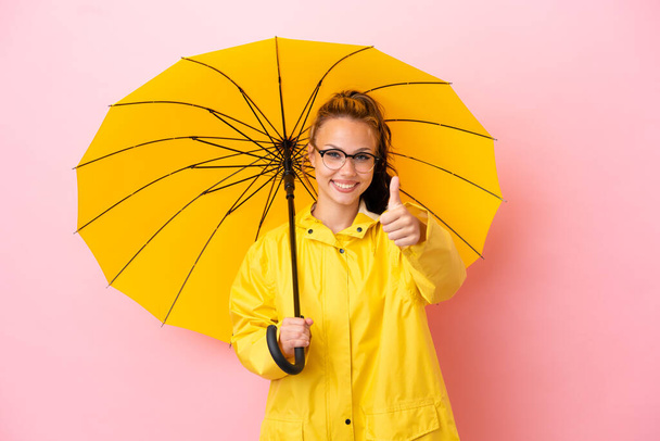 Teenager russische Mädchen mit regendichtem Mantel und Regenschirm isoliert auf rosa Hintergrund mit erhobenen Daumen, weil etwas Gutes passiert ist - Foto, Bild