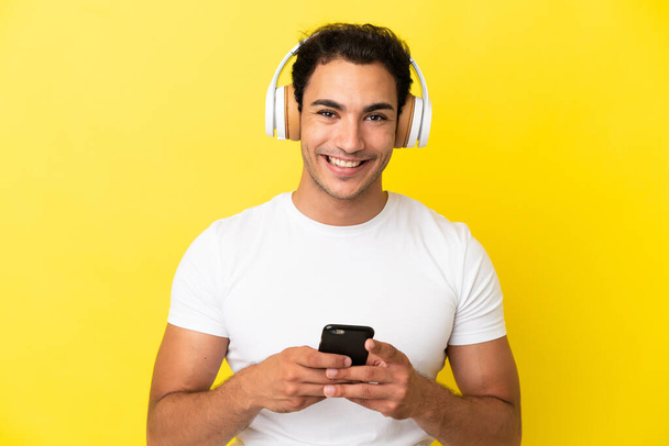 Homem bonito caucasiano sobre fundo amarelo isolado ouvindo música com um celular e olhando para a frente - Foto, Imagem